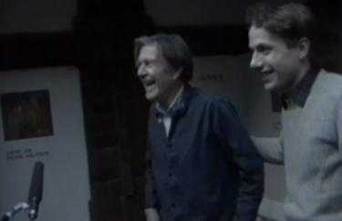 John Cage: Mushroom et Variationes, 1985. Videostill  Zwei Männer im Profil lachen, links ist John Cage, rechts ist Axel Wirths, der John Cage dabei den Arm auf den Rücken legt.